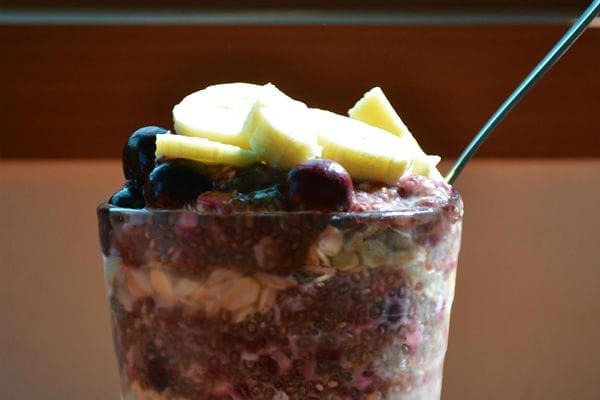 A vibrant vegan breakfast parfait in a clear glass, showing alternating layers of purple blueberry chia seed pudding and light-colored old-fashioned oats, garnished with fresh banana slices on top.