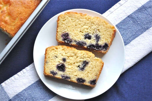A slice of moist vegan blueberry cornbread, studded with vibrant blueberries, resting on a plate, with the golden-brown loaf visible in the background.