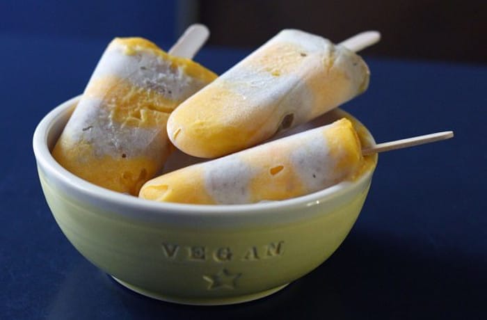 Close-up of vibrant layered mango and banana vegan ice cream pops in clear containers, showing distinct yellow and orange layers with a wooden stick in the center.