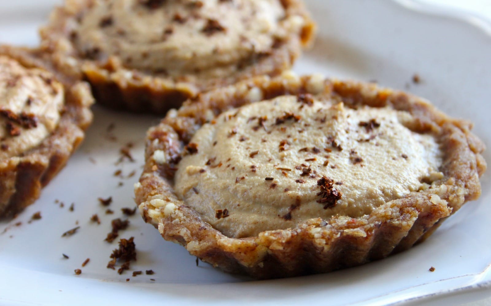 Close-up of a raw vanilla rooibos tartlet, dusted with cacao and cinnamon, served on a small plate, showcasing its creamy filling and nut crust.