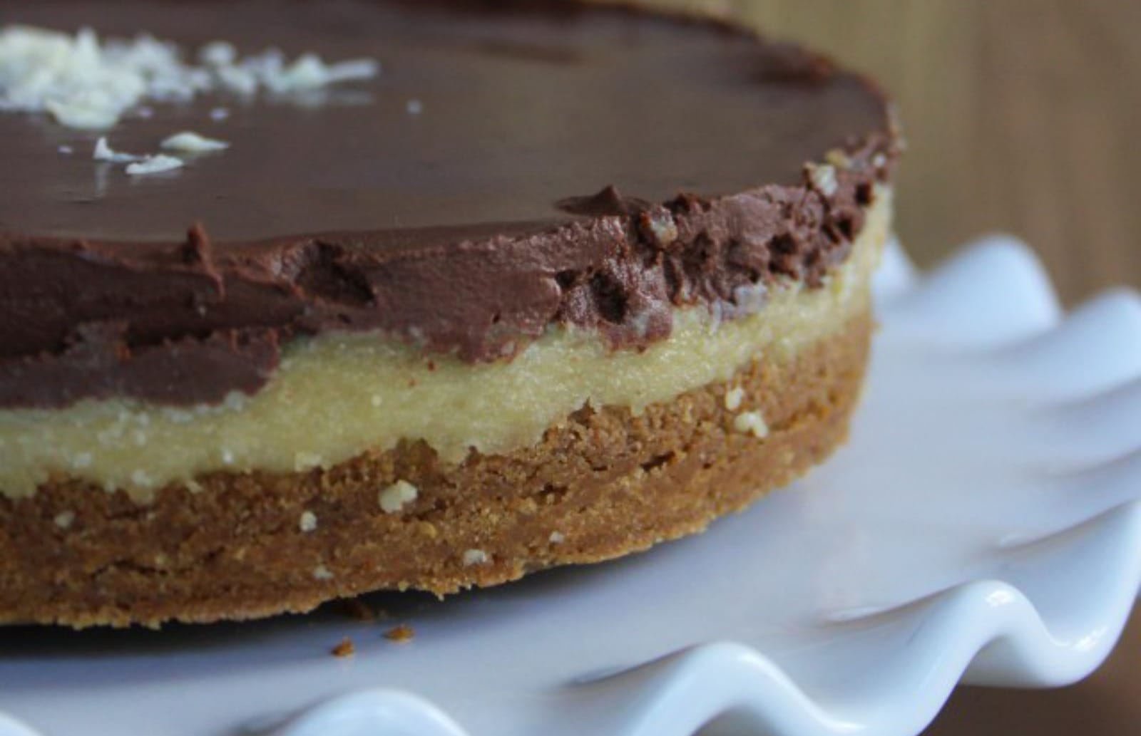 A slice of vegan Irish Cream Chocolate Cheesecake on a white plate, showing the biscuit base, creamy filling with ganache-filled holes, and white chocolate shavings.