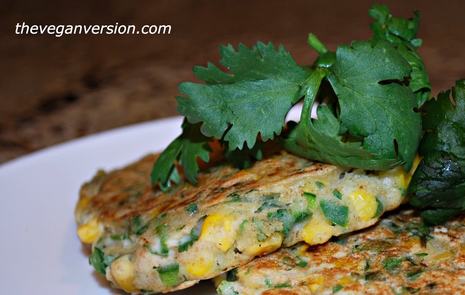 A stack of golden-brown vegan poblano corn pancakes, flecked with green cilantro and red pepper pieces, served on a white plate.