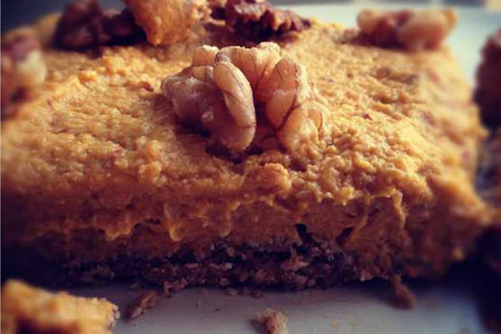 A vibrant slice of no-bake vegan pumpkin pie with a visible textured walnut-oat crust, topped with a smooth, creamy orange filling, garnished on a white plate.