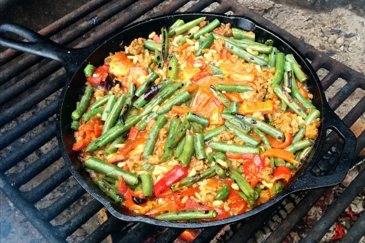 A vibrant Campfire Paella Primavera, served in a paper bowl, showcasing golden rice with smoky socarrat, seitan chorizo, and colorful grilled vegetables.