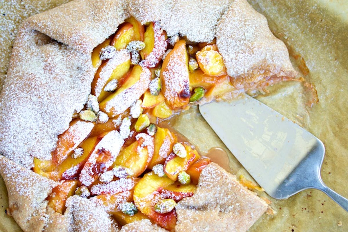 A rustic vegan peach galette with a golden-brown, flaky crust folded over sweet, baked peach wedges, optionally topped with pistachios and powdered sugar.