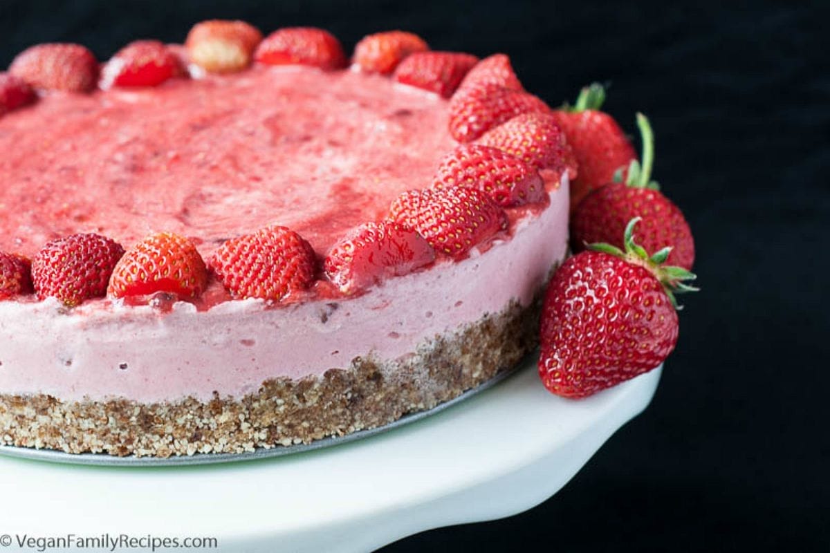 A slice of vibrant No-Bake Strawberry Ice Cream Cake, garnished with fresh halved strawberries, sitting on a plate with the full cake in the background.