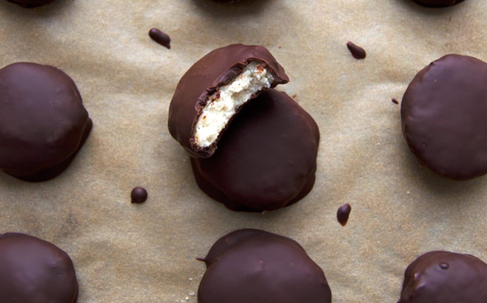 Close-up of several homemade vegan peppermint patties, coated in dark chocolate, some with a slight crack revealing the white minty filling, arranged on a parchment-lined surface.