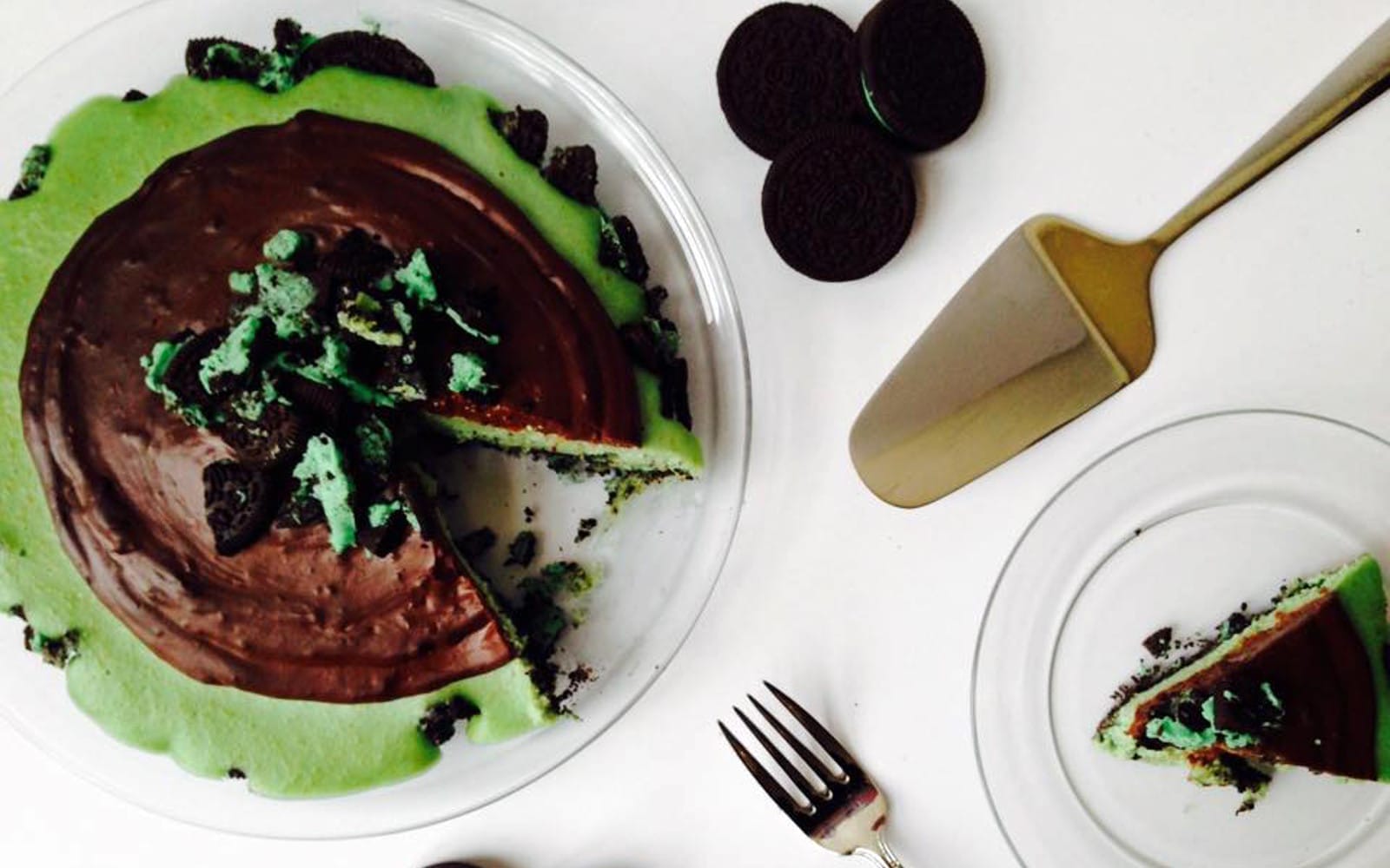 A slice of vibrant green no-bake grasshopper cheesecake, topped with dark chocolate ganache and crumbled mint cookies, on a white plate.