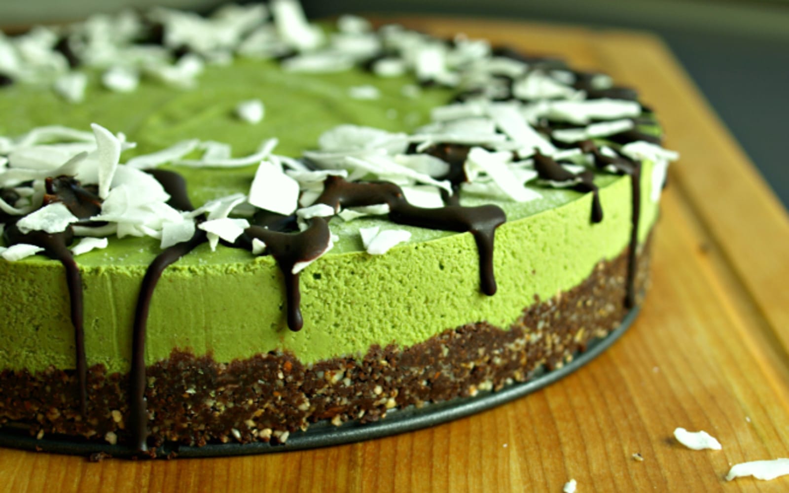 A slice of vibrant green matcha vanilla cheesecake with a dark chocolate cookie crust, drizzled with raw chocolate sauce and sprinkled with coconut shreds, served on a white plate.