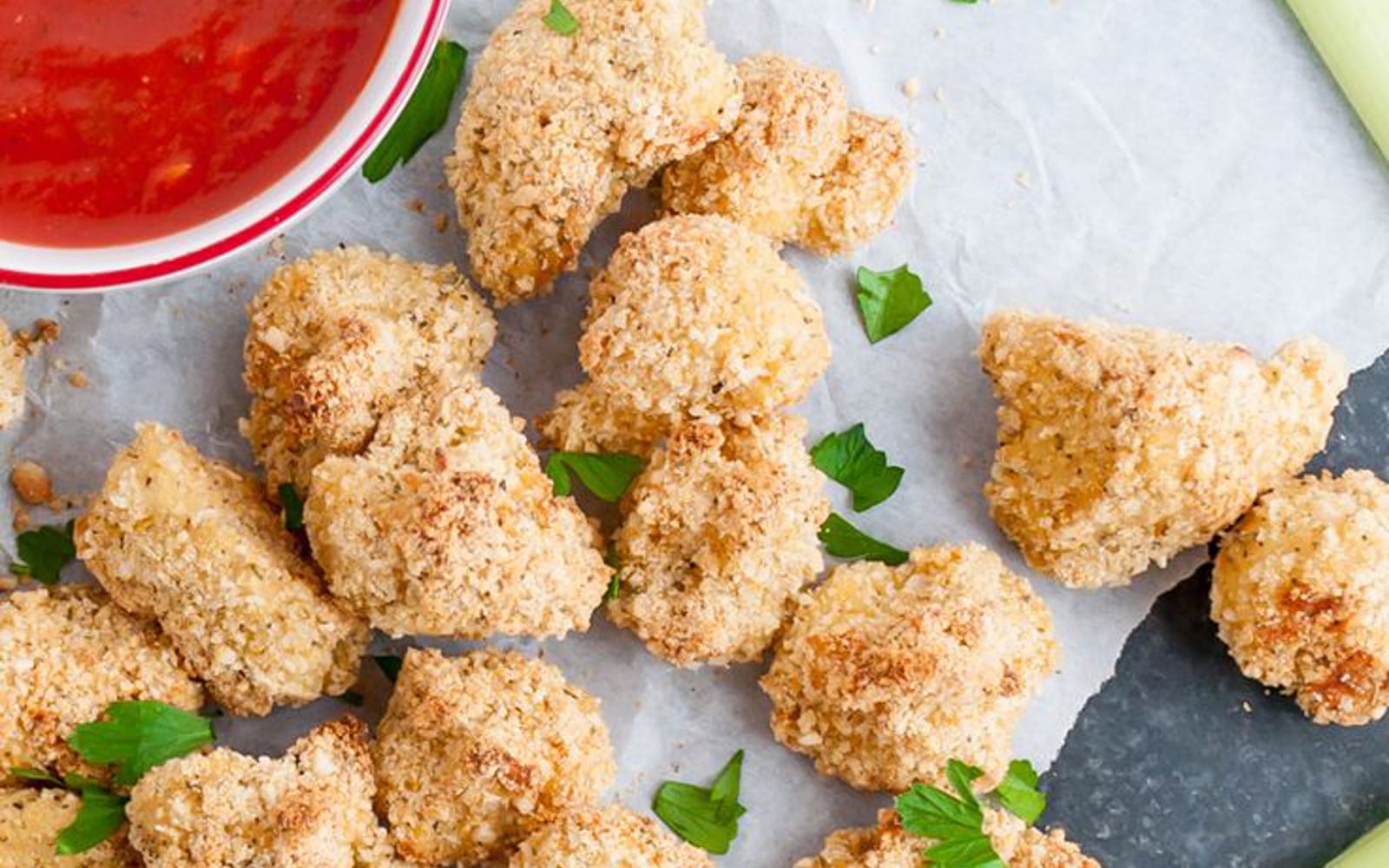 Close-up of golden brown, crispy vegan Parmesan cauliflower bites arranged on a parchment-lined baking sheet, with a small bowl of warm marinara sauce for dipping in the background.