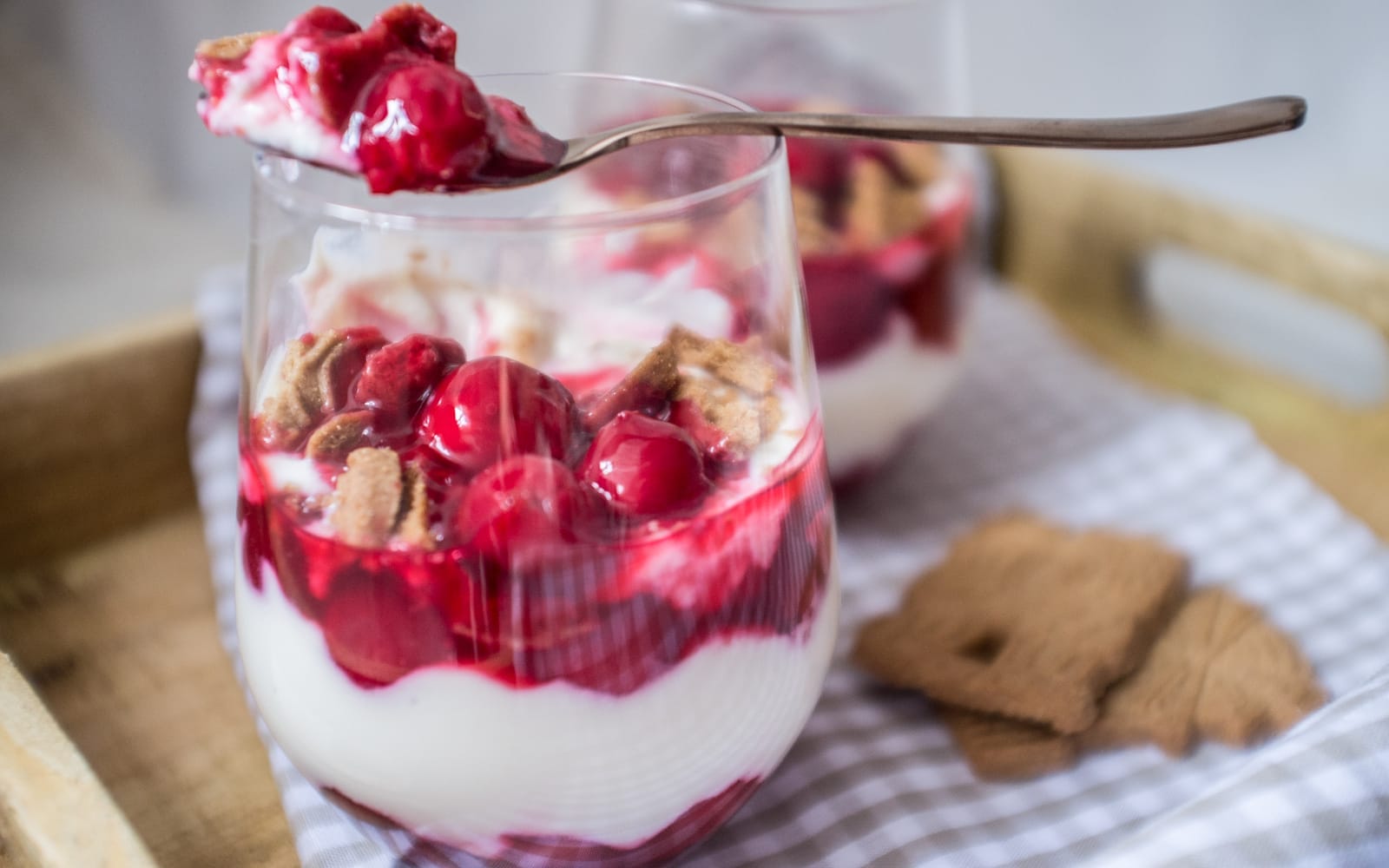 A clear glass jar shows layers of crumbled vegan speculoos cookies, creamy almond yogurt, and bright red cherry compote, topped with more cherries.