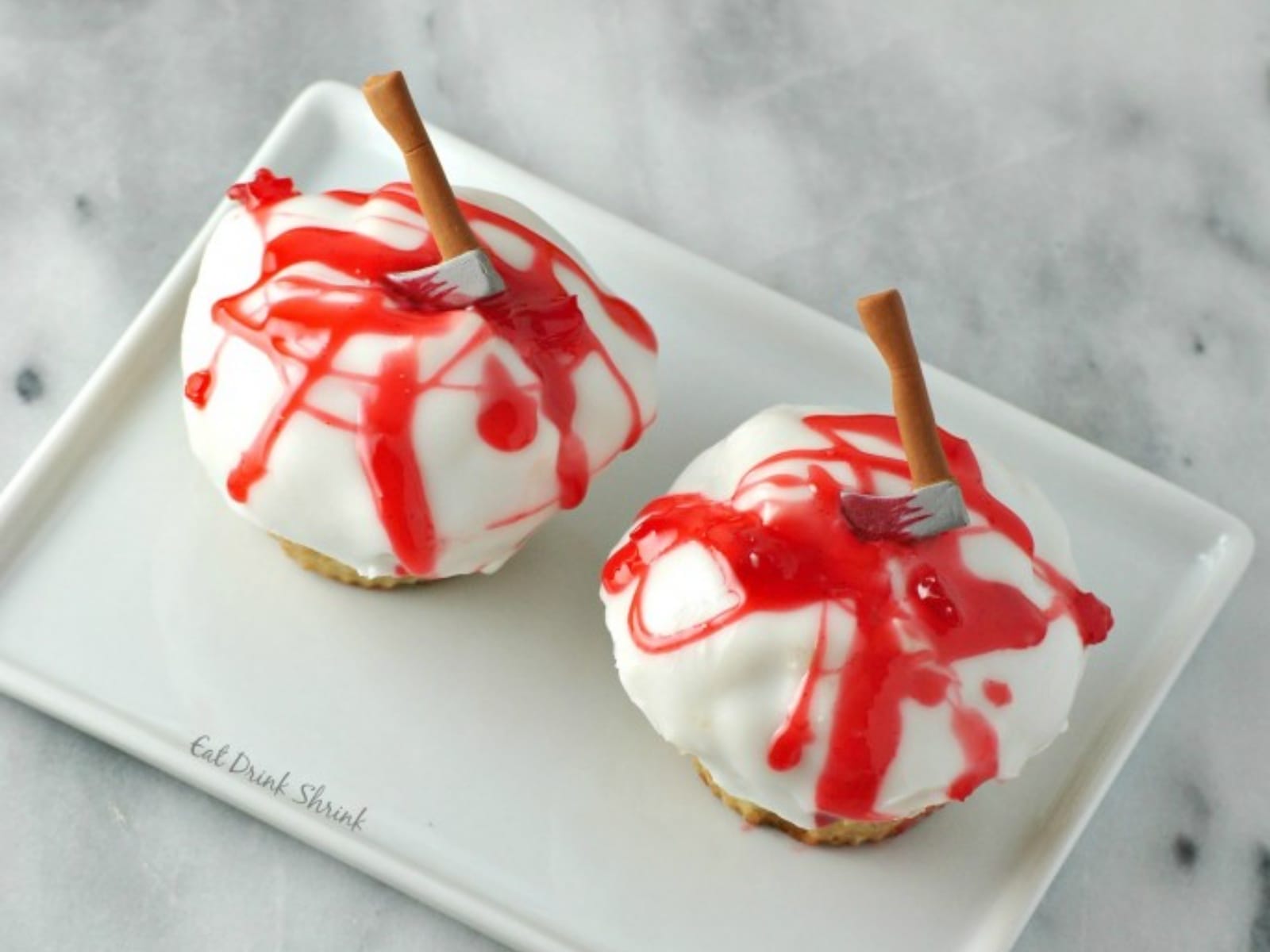 A plate of Bloody Axe Cupcakes, each topped with white icing, a drizzle of red cherry glaze, and a small decorative hatchet, against a dark background.